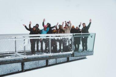 Group of people on a glass observation deck in snowy mountain landscape, waving at the camera. | © manueldietrichphotography