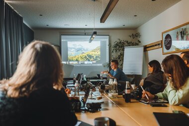 A group of people in a conference room watching a presentation on a screen, with notes and laptops on the table. | © manueldietrichphotography