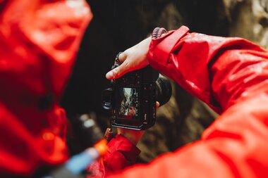 Close-up of a person in red outdoor clothing holding a camera in front of a rocky background. | © manueldietrichphotography