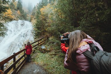 A person photographing a waterfall in autumn forest, surrounded by colorful leaves and trees. | © manueldietrichphotography