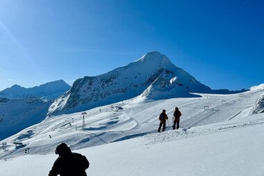 Two people in a snowy landscape with a tall, snow-covered mountain in bright sunlight. | © Zell am See-Kaprun Tourismus