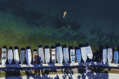 Aerial view of boats on a lakeshore, with a person swimming in the water and a diver above, against a backdrop of water and greenery. | © Zell am See-Kaprun