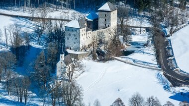 Aerial view of a medieval castle in a snowy winter landscape surrounded by forest and a road. | © Mairitsch 