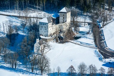 Aerial view of a medieval castle in a snowy winter landscape surrounded by forest and a road. | © Mairitsch 