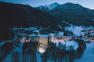 Kaprun Castle at dusk in a snow-covered landscape, surrounded by mountains, illuminated and visible in the valley. | © Zell am See-Kaprun Tourismus