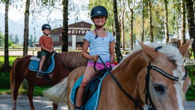 Children riding horses at Seehof with trees in the background and a building in the woods. | © Zell am See-Kaprun Tourismus