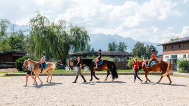 Children learning to ride horses outdoors at a riding school with mountains in the background. | © Zell am See-Kaprun Tourismus