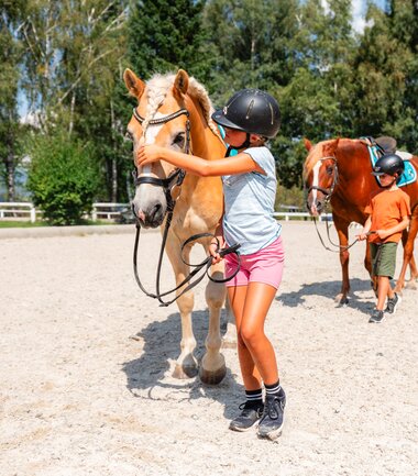 Children engaging with horses during a fun program at an equestrian center with trees in the background. | © Zell am See-Kaprun Tourismus