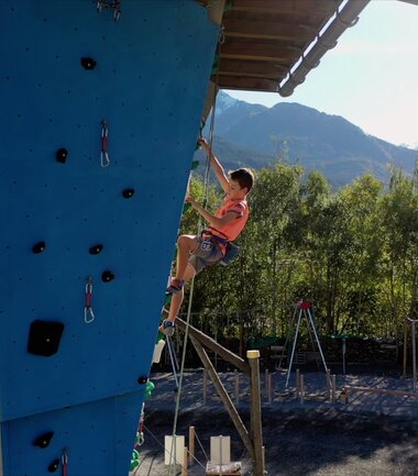 Young person climbing an outdoor climbing wall with mountains in the background. | © Zell am See-Kaprun Tourismus