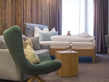 Cozy hotel room with a double bed, a comfortable armchair, a small table with a water bottle, and curtains at the window. | © Nikolaus Faistauer Photograhpy