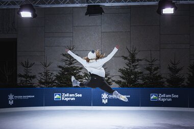 Figure skater jumps during a performance on an indoor ice rink, with Christmas trees in the background. | © Zell am See-Kaprun Tourismus