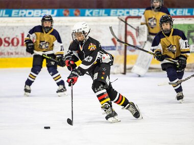 Ice hockey player with helmet and gear playing on the ice, surrounded by other players and a goal. | © Johannes Radlwimmer