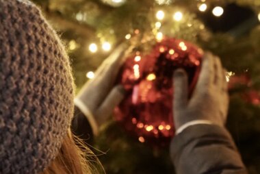 Person decorating a Christmas tree with a large red bauble surrounded by string lights. | © Zell am See-Kaprun Tourismus
