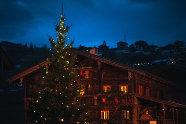 Wooden house decorated for Christmas with a lit Christmas tree in front of a dark sky in Kaprun. | © Johannes Radlwimmer