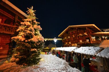 Christmas market at night in Kaprun with a snow-covered tree and festive wooden stalls lit up. | © Zell am See-Kaprun Tourismus