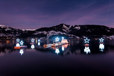 Illuminated stars on a lake reflect in the water, with snow-covered mountains in the background at dusk. | © Christian Mairitsch