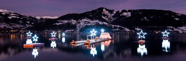 Illuminated stars on a lake reflect in the water, with snow-covered mountains in the background at dusk. | © Christian Mairitsch