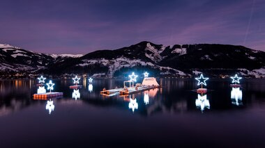 Illuminated stars on a lake reflect in the water, with snow-covered mountains in the background at dusk. | © Christian Mairitsch