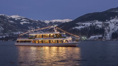 A illuminated boat on a lake at night with snow-covered mountains in the background. | © Schmittenhöhe