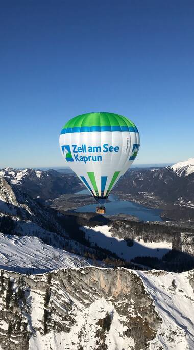 Hot air balloon with 'Zell am See Kaprun' lettering flying over snowy mountains under clear sky. | © Zell am See-Kaprun Tourismus