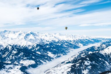 Two hot air balloons flying over snow-covered mountains in a clear sky scene. | © Zell am See-Kaprun Tourismus