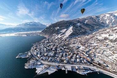 Aerial view of Zell am See with snow-covered town, mountains in the background, and hot air balloons in the sky. | © Jürgen Feichter