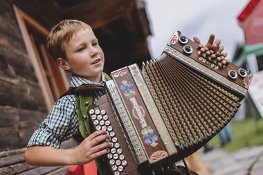 Child playing an accordion at a culinary market in Kaprun, with a wooden house in the background. | © Oberhauser Photography