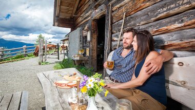 Two people sit at a wooden table outside a mountain cabin, enjoying drinks and a picnic with a flower arrangement in a rural area. | © Bernhard Moser Photography