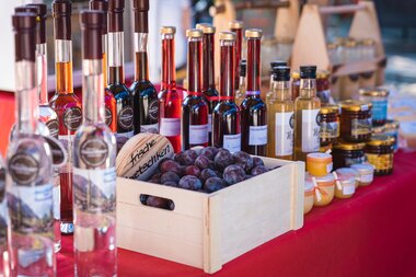 Various bottles of liqueur or juice, a wooden box with plums, honey jars, and other products on a market stall table. | © Zell am See-Kaprun Tourismus