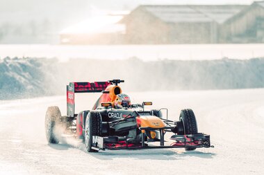 Formula 1 car racing on a snowy track, surrounded by snow-covered hills and buildings in the background. | © GP ICE RACE, Max Zappolino