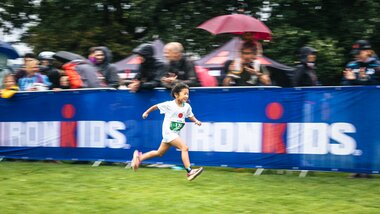 A child running happily at the IRONKIDS race in Zell am See-Kaprun with spectators holding umbrellas in the background. | © Zell am See-Kaprun, JOE