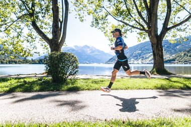 A runner jogging along a lake with mountains in the background, surrounded by trees and greenery. | © Zell am See-Kaprun, JOE