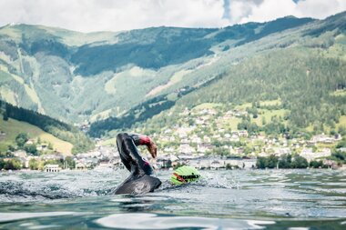 Triathlete swimming in Lake Zell with mountains and a town in the background. | © Zell am See-Kaprun Tourismus