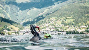 Triathlete swimming in Lake Zell with mountains and a town in the background. | © Zell am See-Kaprun Tourismus