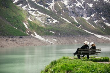 Two people sit on a bench by the lake shore surrounded by mountains with patches of snow. | © Michael Freudenthaler
