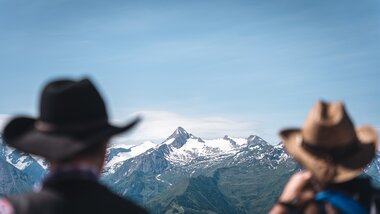 Two people with hats in the foreground, overlooking the Alps with snow-capped peaks in the background. | © Zell am See-Kaprun Tourismus