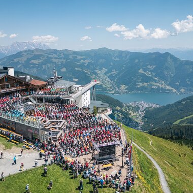 Large outdoor event on a mountain terrace with views of mountains and a lake, many visitors, and an outdoor stage. | © Zell am See-Kaprun, JOE