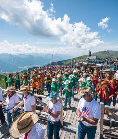 Large line dance event with many participants in traditional costumes and hats against an alpine mountain backdrop on a sunny day. | © Zell am See-Kaprun, JOE