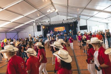People line dancing at AlpFestival 2025 inside a large tent, with a stage in the background. | © Zell am See-Kaprun, JOE