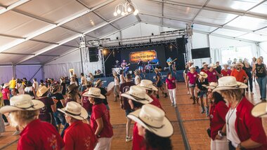 People line dancing at AlpFestival 2025 inside a large tent, with a stage in the background. | © Zell am See-Kaprun, JOE