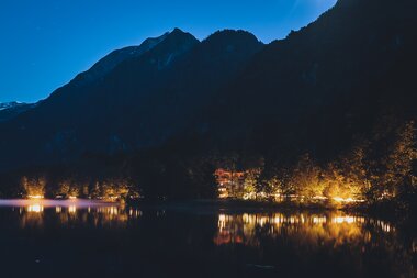 Night view over water with reflections of lights from trees and buildings on the shore, against mountains in darkness. | © EXPA, Jürgen Feichter