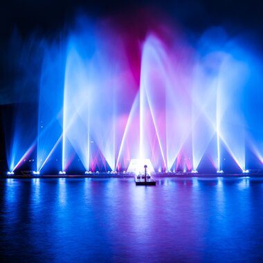 Colorful water light display with vibrant water jets rising on a lake at night, reflecting on the water. | © Christian Mairtisch