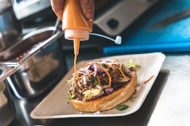 Close-up of a sandwich being topped with sauce from a squeeze bottle, on a wooden serving plate. | © Zell am See-Kaprun Tourismus