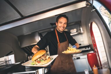 A friendly chef presents a freshly prepared burger in a mobile kitchen at a summer night festival. | © Zell am See-Kaprun Tourismus