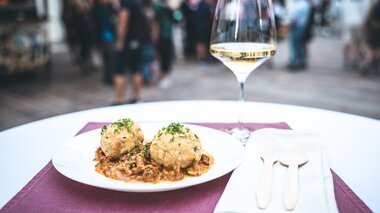 Elegant table setting with a glass of white wine and a plate with dumplings in an outdoor summer festival setting, with a crowd in the background. | © Zell am See-Kaprun Tourismus