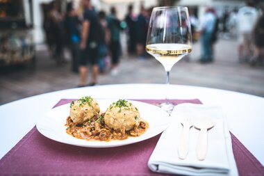 Elegant table setting with a glass of white wine and a plate with dumplings in an outdoor summer festival setting, with a crowd in the background. | © Zell am See-Kaprun Tourismus