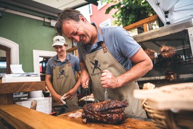 Person grilling a large piece of meat at a summer night festival, smiling. | © Zell am See-Kaprun Tourismus