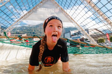 Child participating in the Spartan Kids Race, crossing a water obstacle under a large metal roof with mountains and sky in the background. | © Zell am See-Kaprun Tourismus