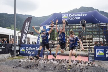 Participants in a Spartan Race crossing a muddy, uneven surface outdoors with mountains in the background. | © Zell am See-Kaprun Tourismus