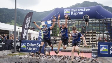 Participants in a Spartan Race crossing a muddy, uneven surface outdoors with mountains in the background. | © Zell am See-Kaprun Tourismus